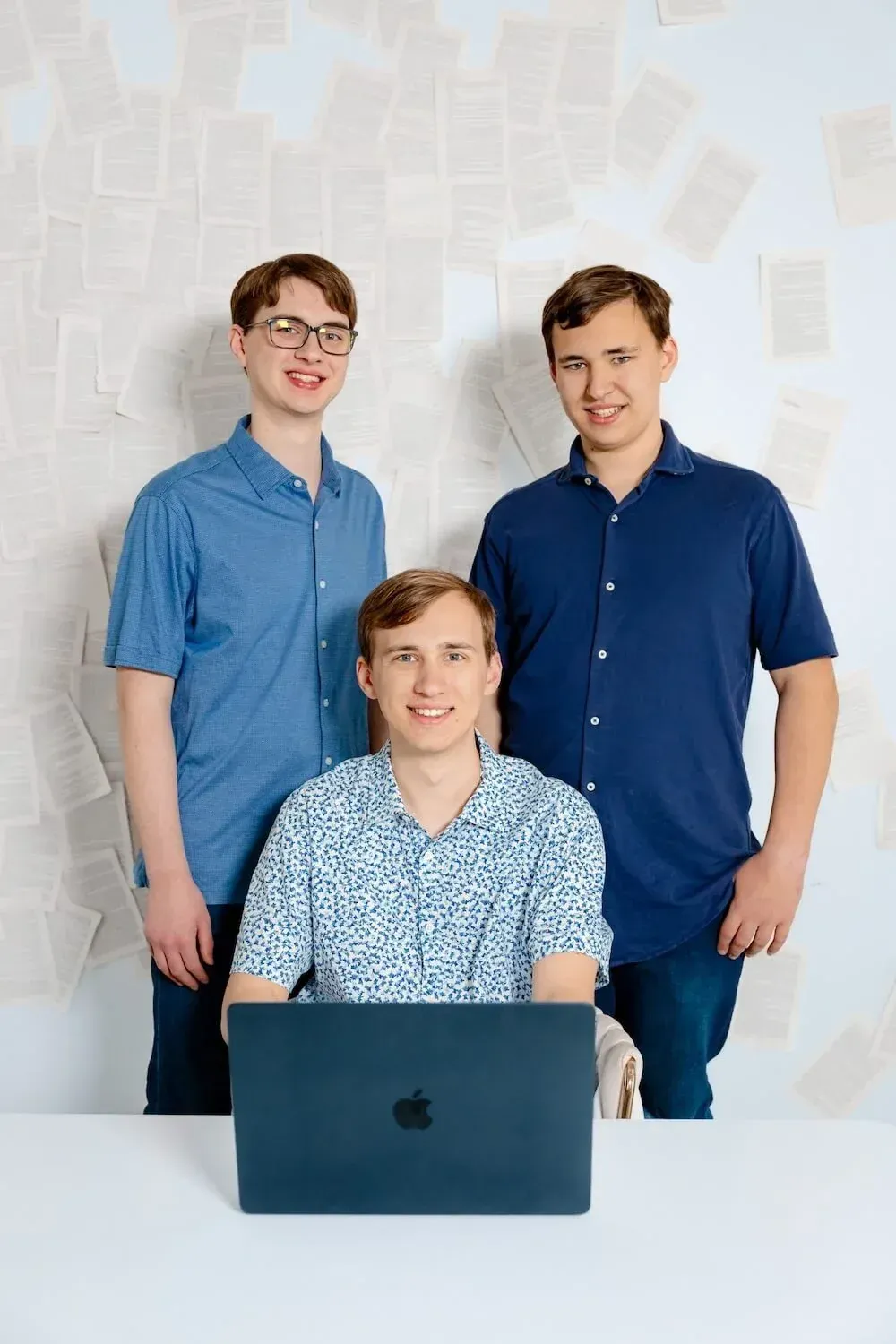 Three young men, two standing and one sitting at a desk, in a room with a wall covered in scattered pages, smiling at the camera.